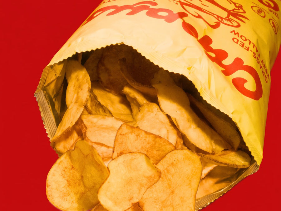 An open bag of Chippys spilling golden kettle chips on a red background